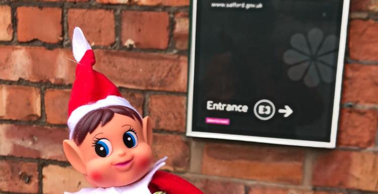 A picture of Irwell the Healthwatch Salford elf, dressed in a red hat and suit, standing outside a sign that reads 'Eccles Town Hall'