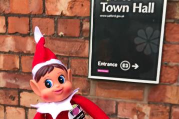 A picture of Irwell the Healthwatch Salford elf, dressed in a red hat and suit, standing outside a sign that reads 'Eccles Town Hall'